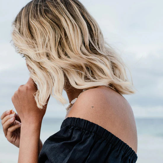 Woman with wavy blonde hair touching her face against a blurred natural background while wearing the Barriere biotin patch with the black diamond symbol on her shoulder