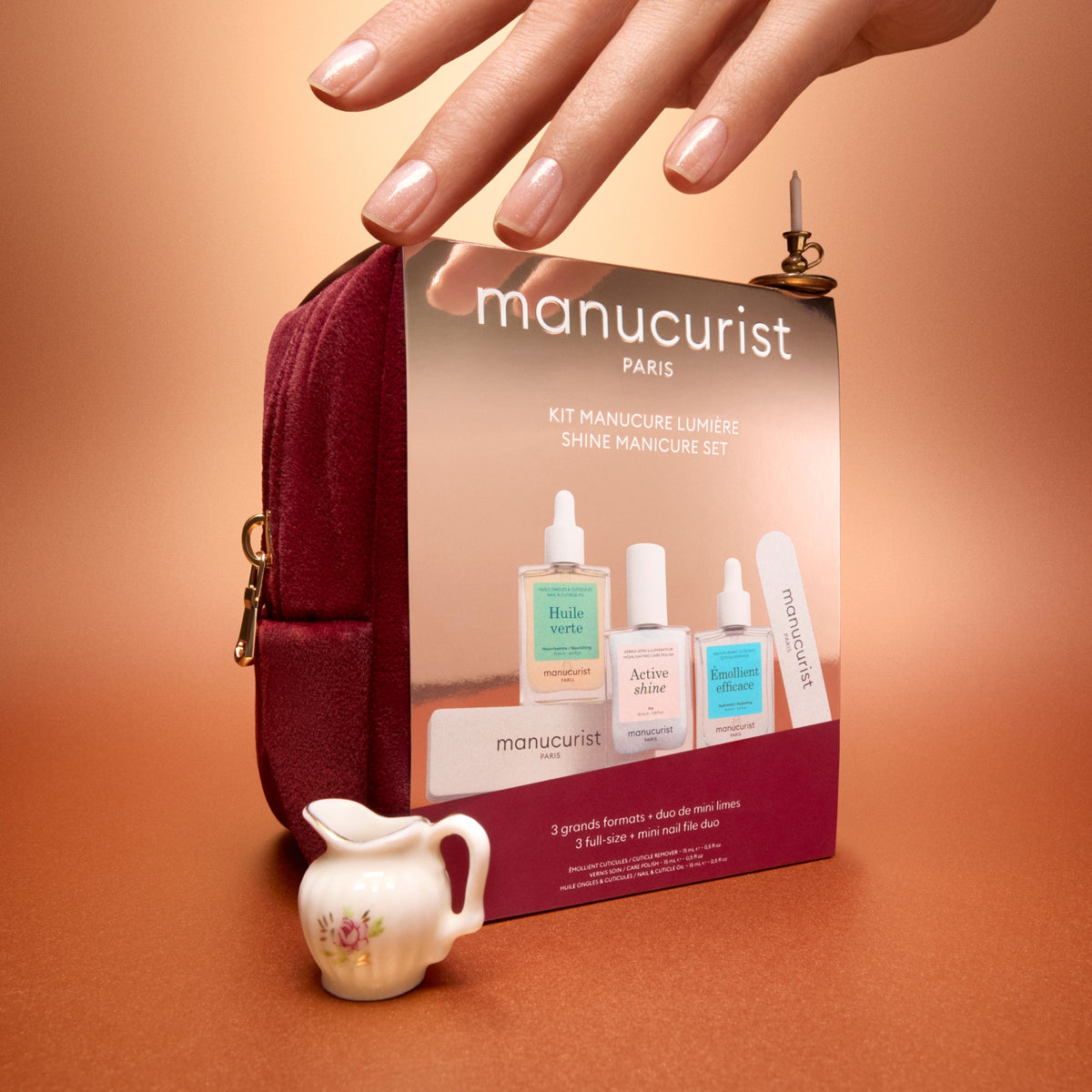 A manicure kit set by Manucurist Paris, featuring a burgundy zippered pouch, a miniature white floral pitcher, and three nail care bottles, arranged on a warm brown surface with a hand resting on top of the pouch.