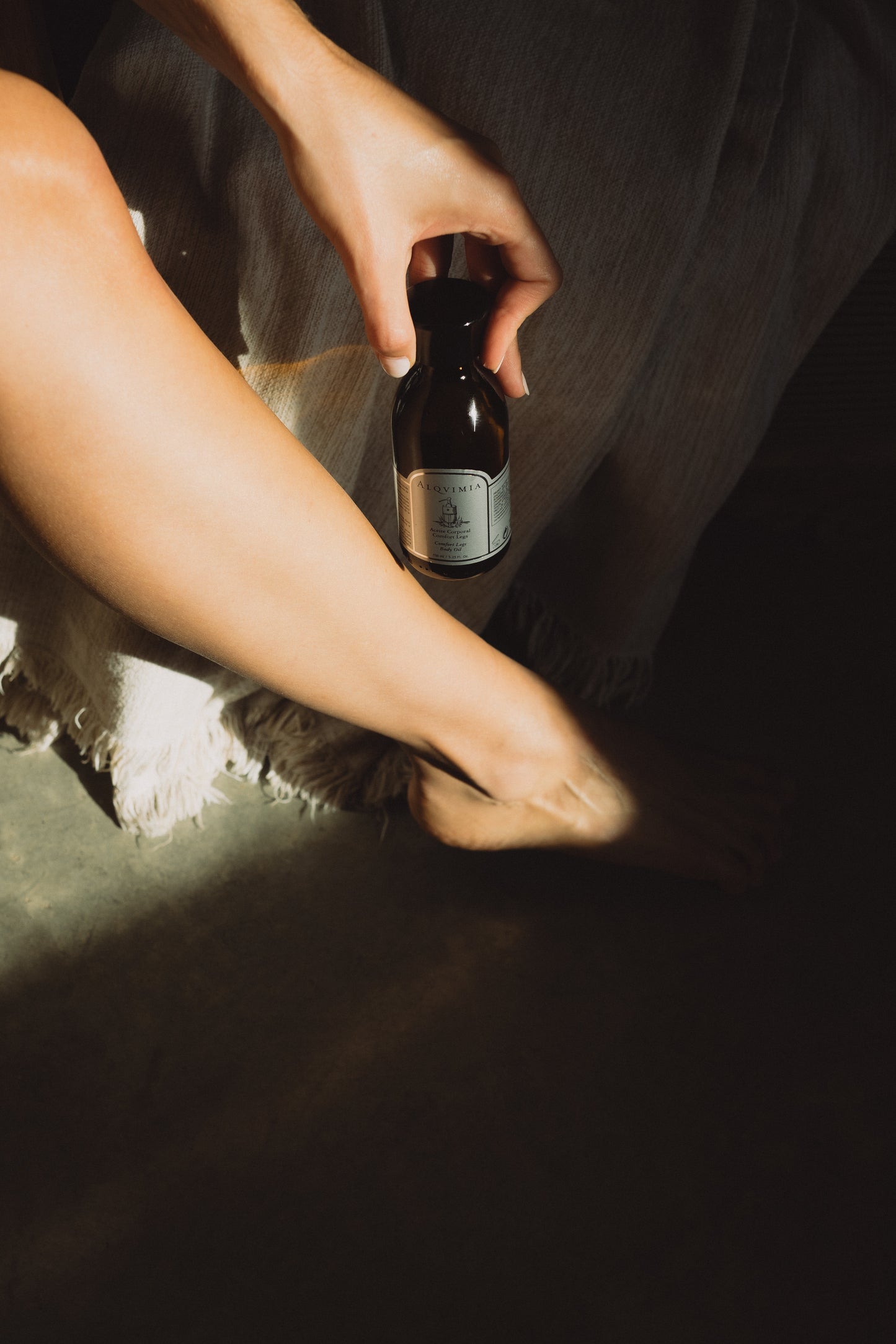A person sitting with legs crossed applies Alqvimia Comfort Legs Body Oil, holding the brown glass bottle near their leg in warm, natural light.