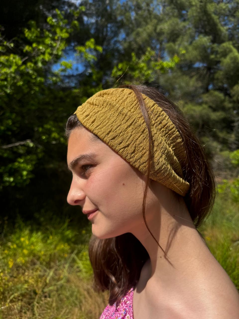 Woman wearing a mustard yellow knitted headband outdoors with greenery in the background