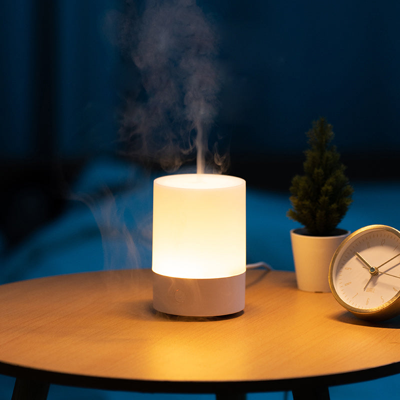 White essential oils diffuser emitting steam on a wooden table with a plant and clock in the background