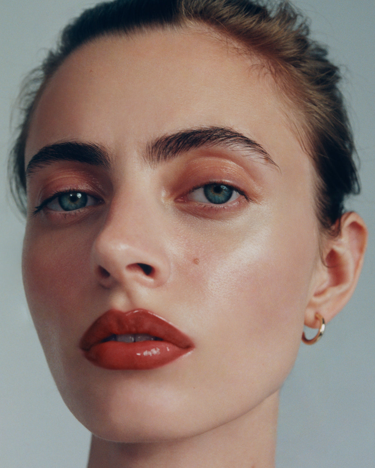 Close-up portrait of a woman with blue eyes wearing a small gold hoop earring and and the Manasi 7 tinted beauty potion in the shade kagero  against a light gray background.