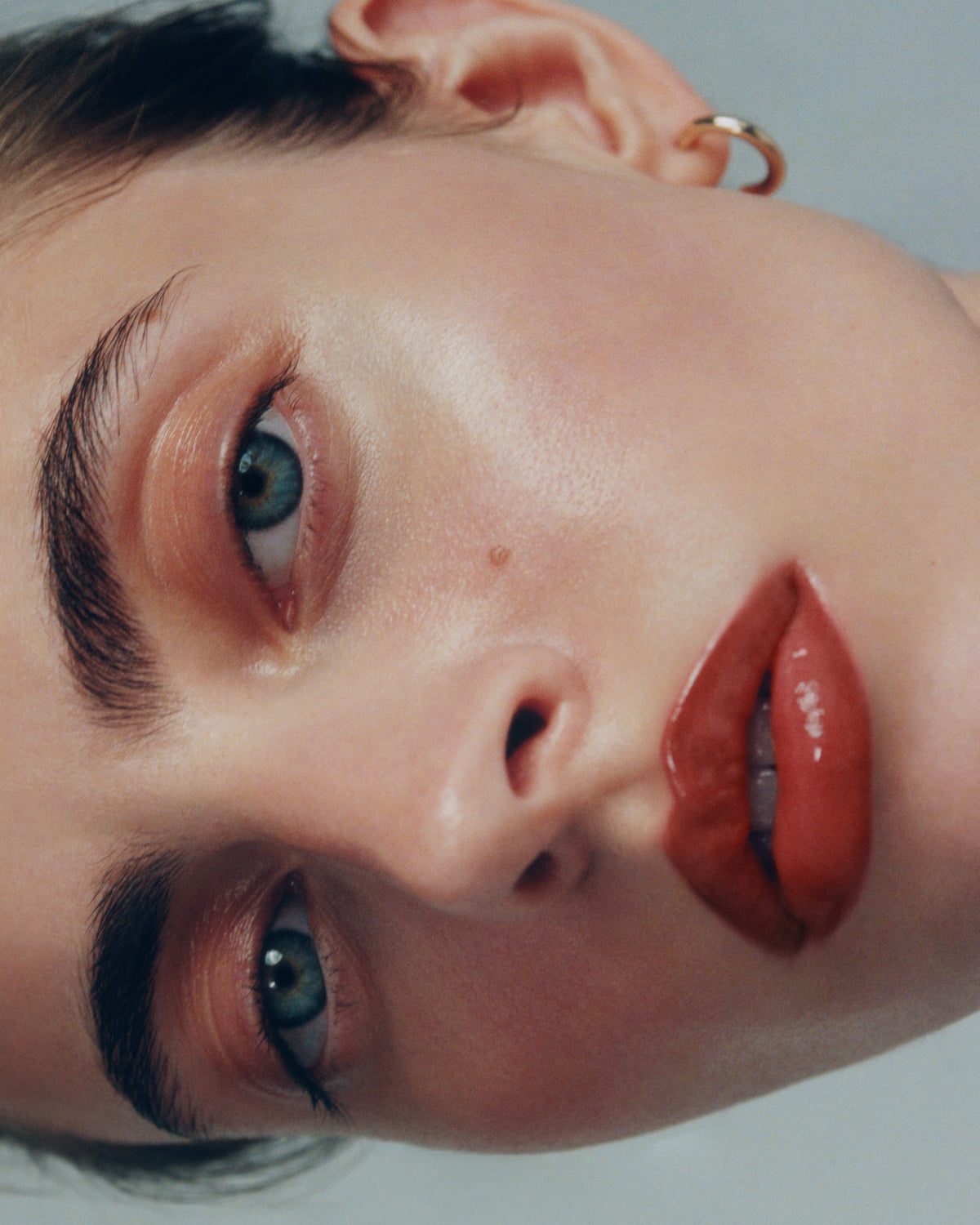 Close-up portrait of a woman with blue eyes wearing a small gold hoop earring and and the Manasi 7 tinted beauty potion in the shade kagero  against a light gray background.
