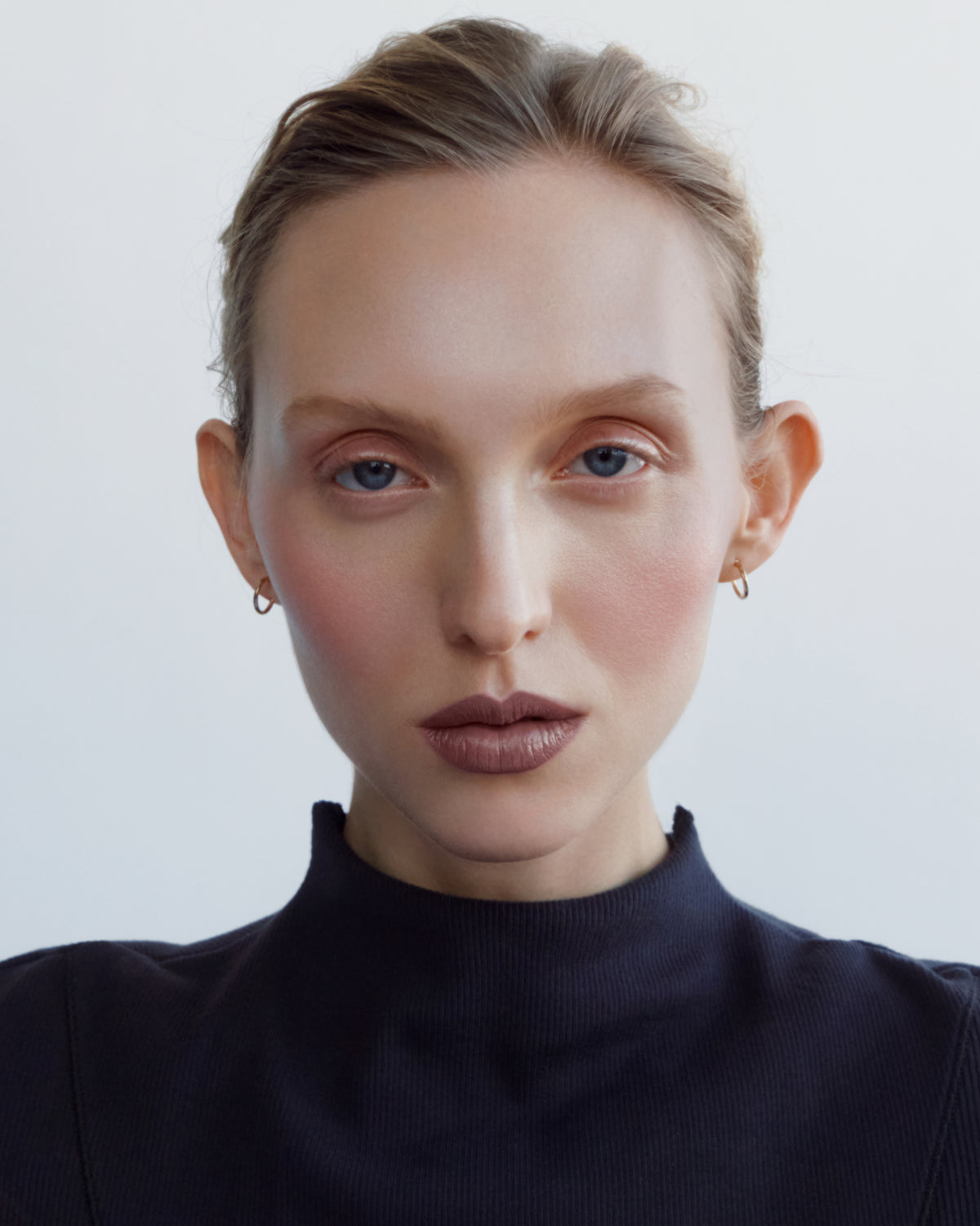 A close-up portrait of a woman with blonde hair pulled back, wearing small gold hoop earrings, a dark navy turtleneck top and the the Manasi 7 tinted beauty potion in the shade simonkai against a light gray background.