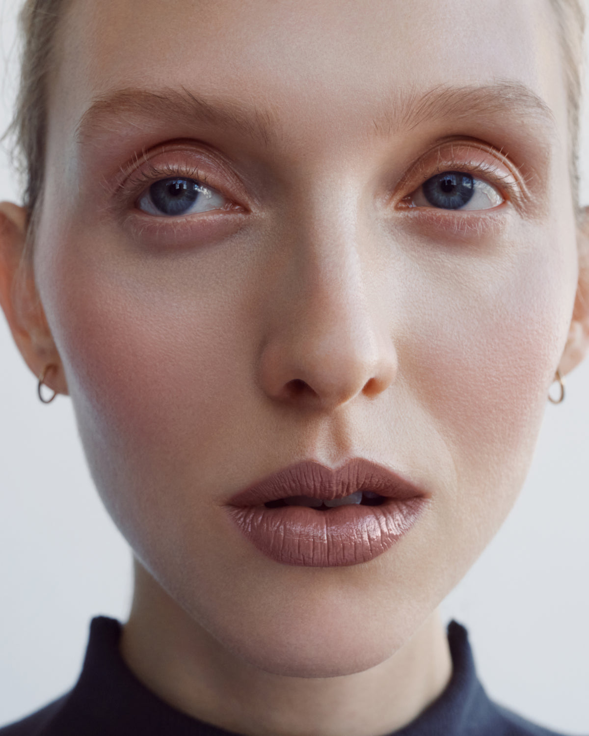 Close-up portrait of a woman with blue eyes wearing a small gold hoop earring and and the Manasi 7 tinted beauty potion in the shade simonkai  against a light gray background.