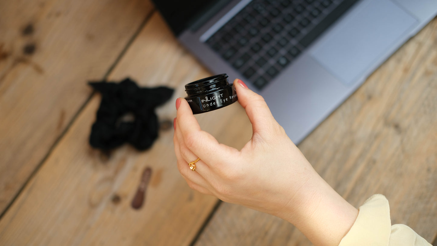 Hand holding a small black container of the Inlight Under eye revive in front of a laptop on a wooden desk