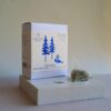 A square white product box with a minimalist blue pine tree illustration, displayed on a white pedestal against a warm beige background. It contains greek herbal blend, tea.  