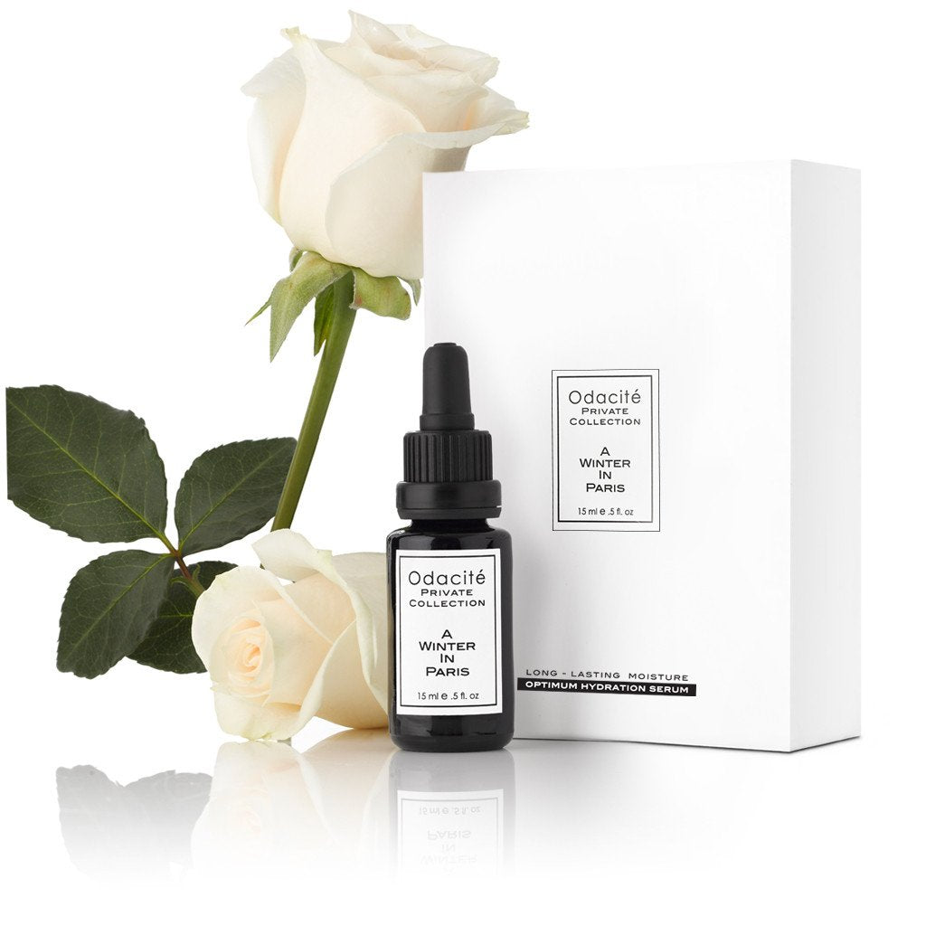 Black dropper bottle of odacite a winter in paris serum with white label next to a white rose and packaging on a white background