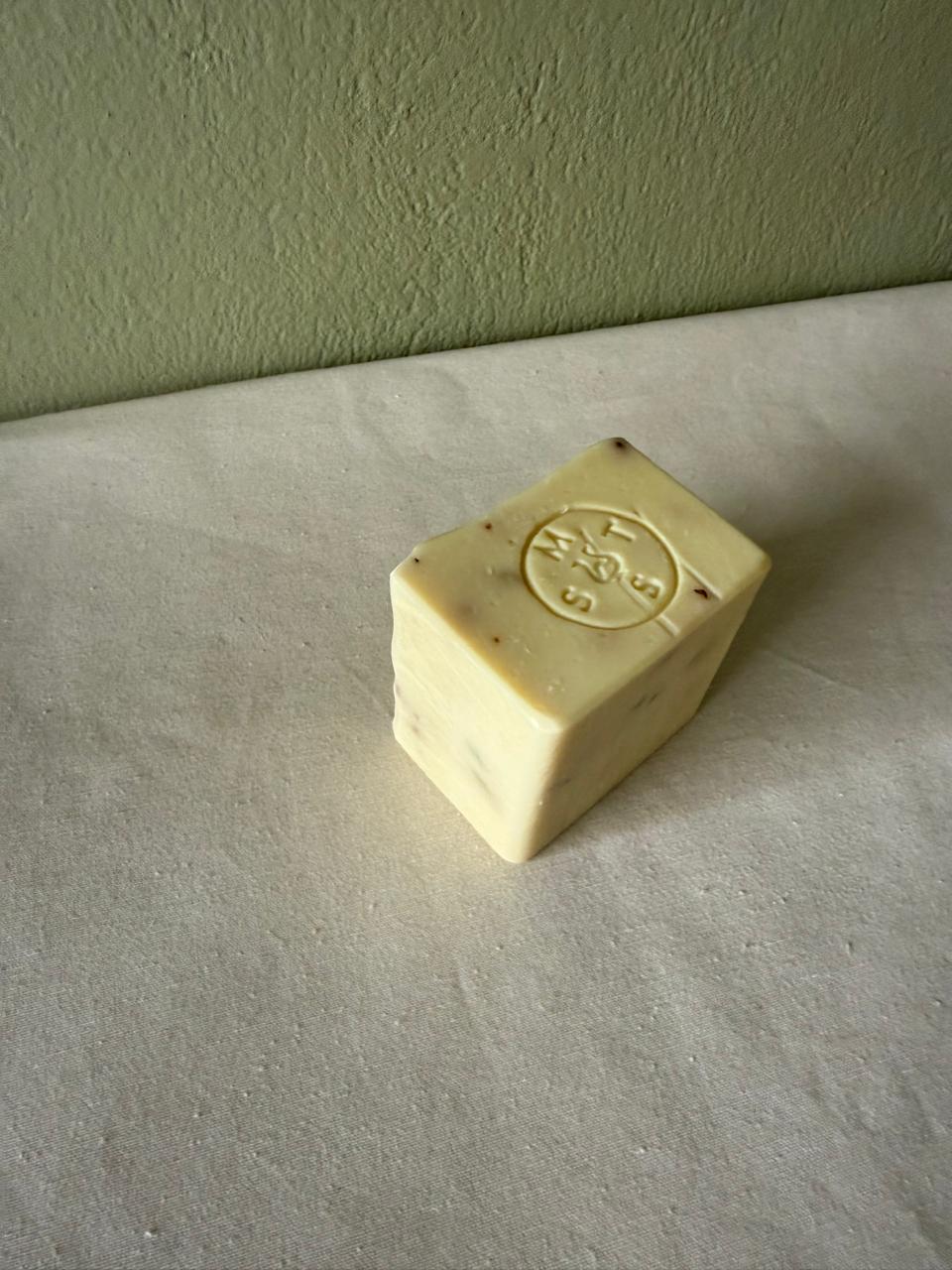 Square bar of lavender soap with a Serendipity Muses logo on a white surface and green wall background