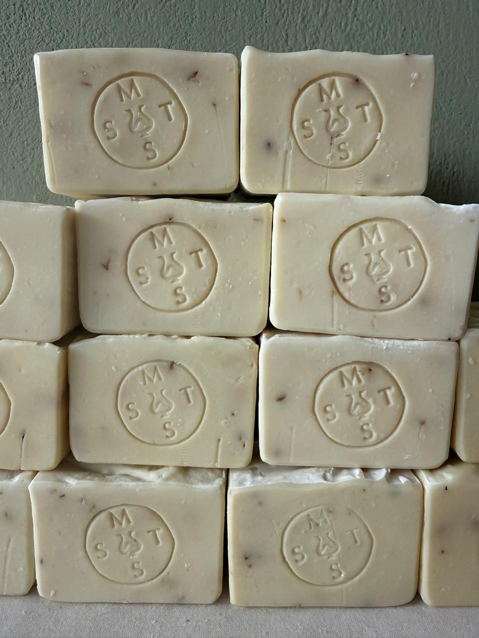 Close up of a stack of beige lavender soap bars with the Serendipity Muses logo on a white surface and green background