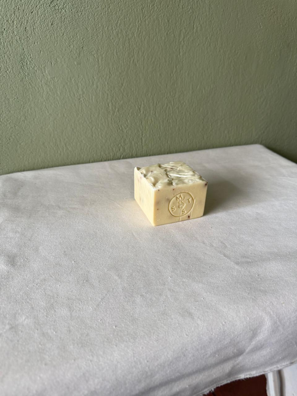 Square bar of lavender soap with a Serendipity Muses logo on a white surface and green wall background