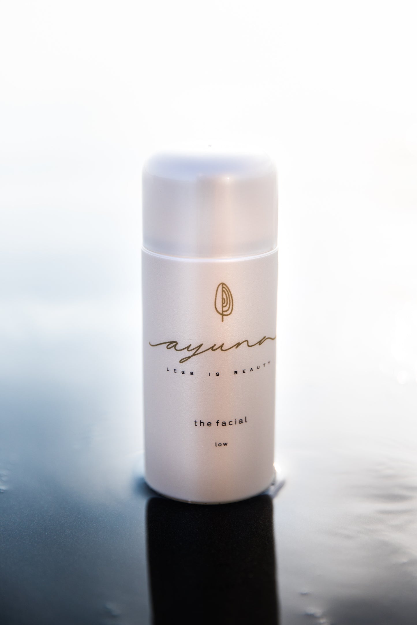 Skincare bottle labeled 'ayuna the facial - low' on a reflective surface with a white background