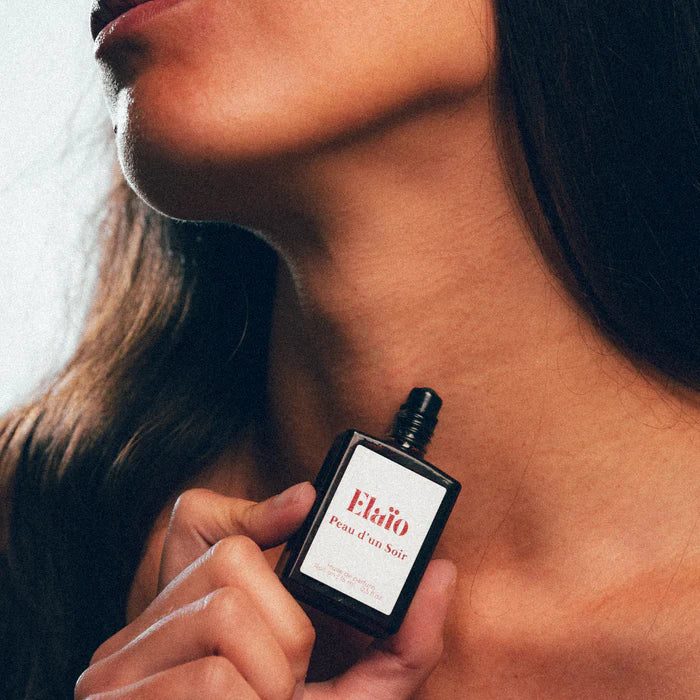 a woman holding a Perfume bottle labeled 'Elaïo Peau d'un Soir' close to her neck