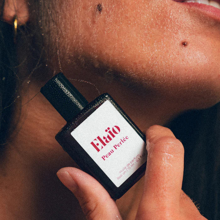 Person holding a sElaéo Peau Perlée perfume bottle close to their neck.