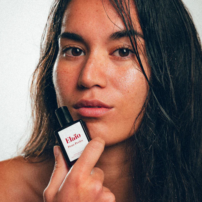 Woman holding a Elaéo Peau Perlée perfume bottle close to her face against a neutral background