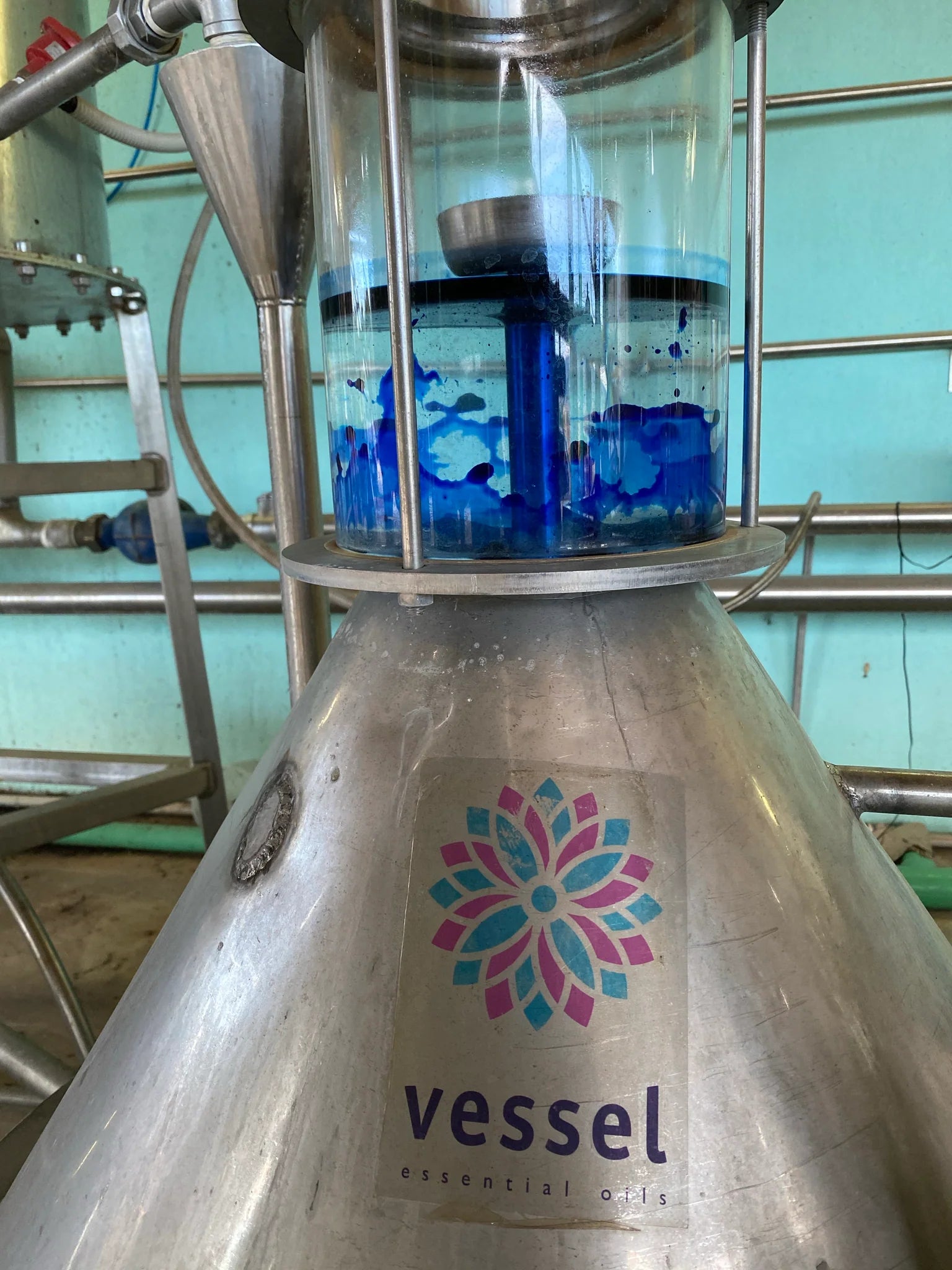 Large metal tank with 'Vessel' logo and blue chamomile essential oil inside, on a teal background.