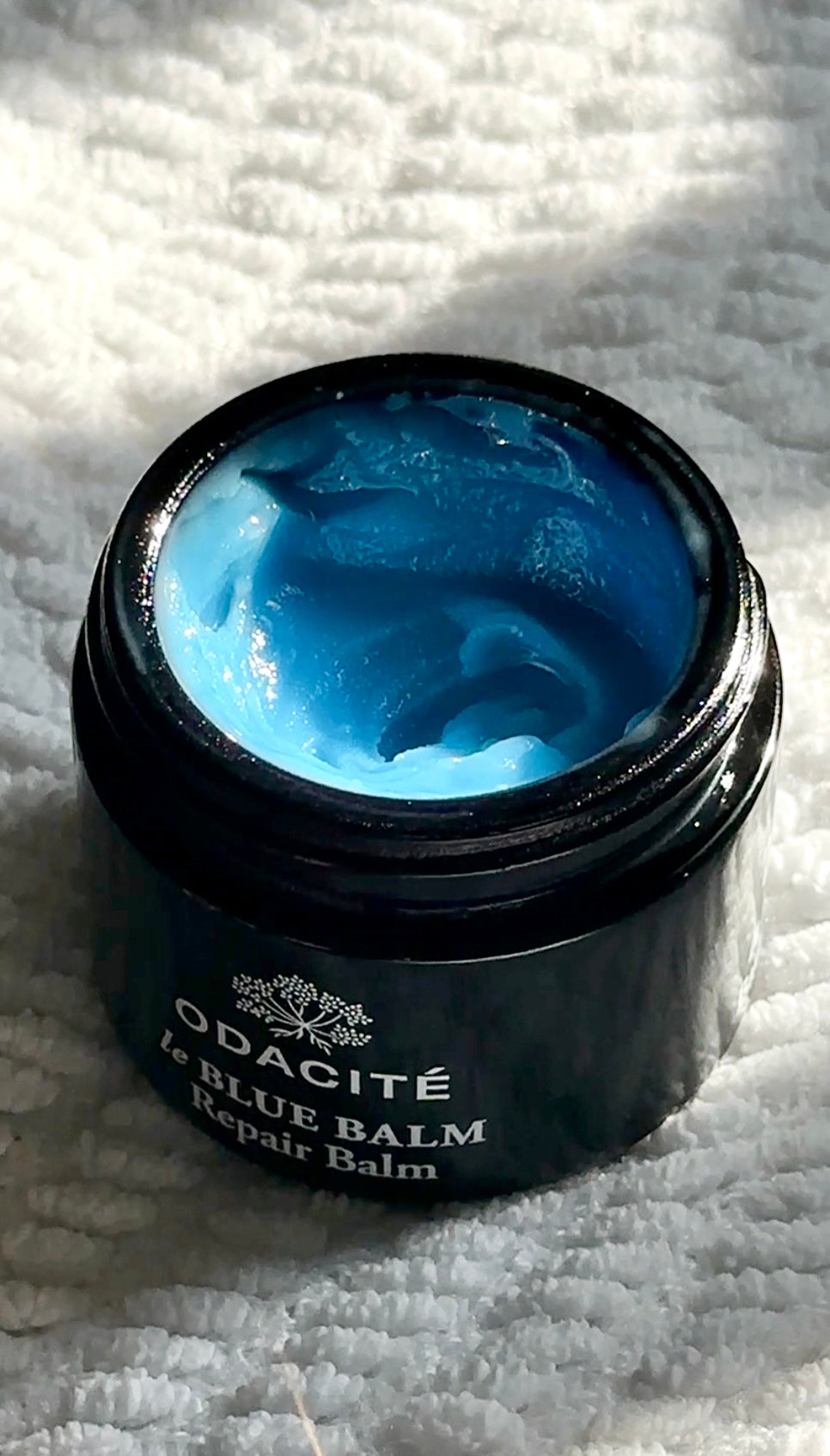 Black jar of blue balm labeled 'Odacite Le Blue Balm' on a textured white surface