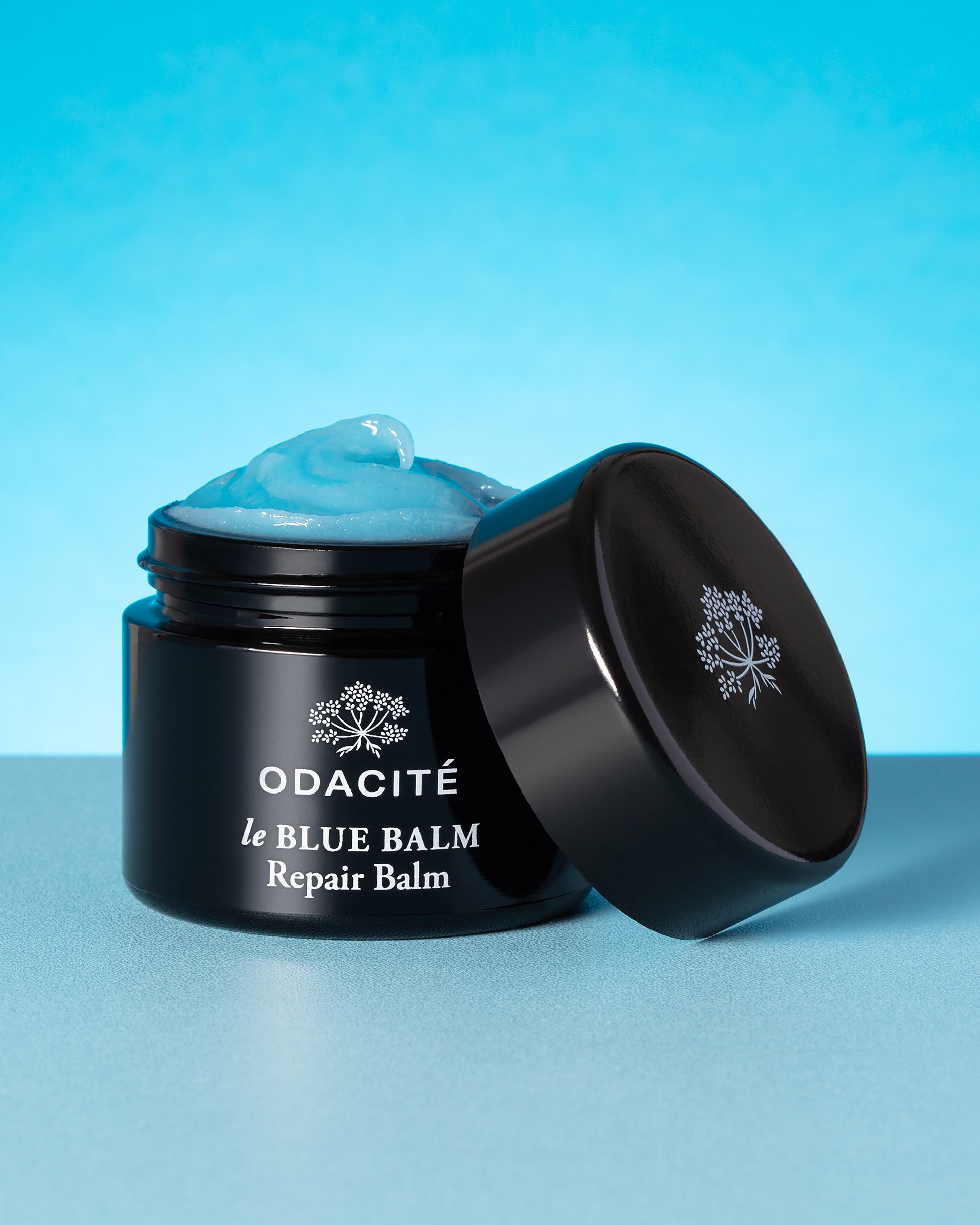 Black jar of Odacite le Blue Balm Repair Balm with blue cream on a blue background