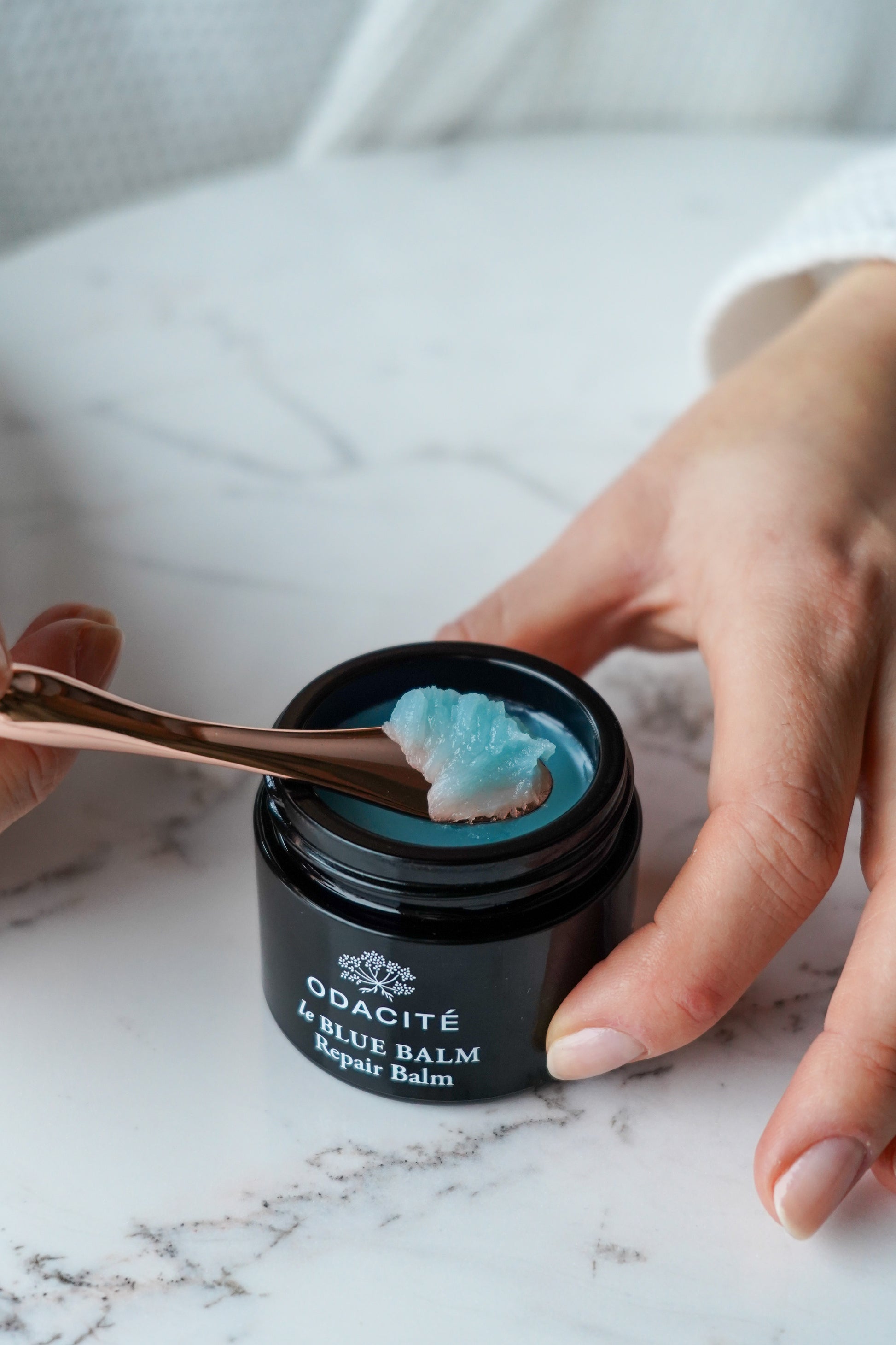 Person using a spoon to take blue balm from a jar labeled 'Odaciite le Blue Balm' on a marble surface.