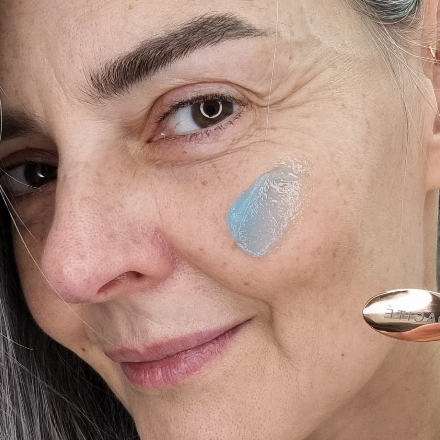 Close-up of a woman's face with a le Blue Balm by Odacite on her cheek and a dropper above.
