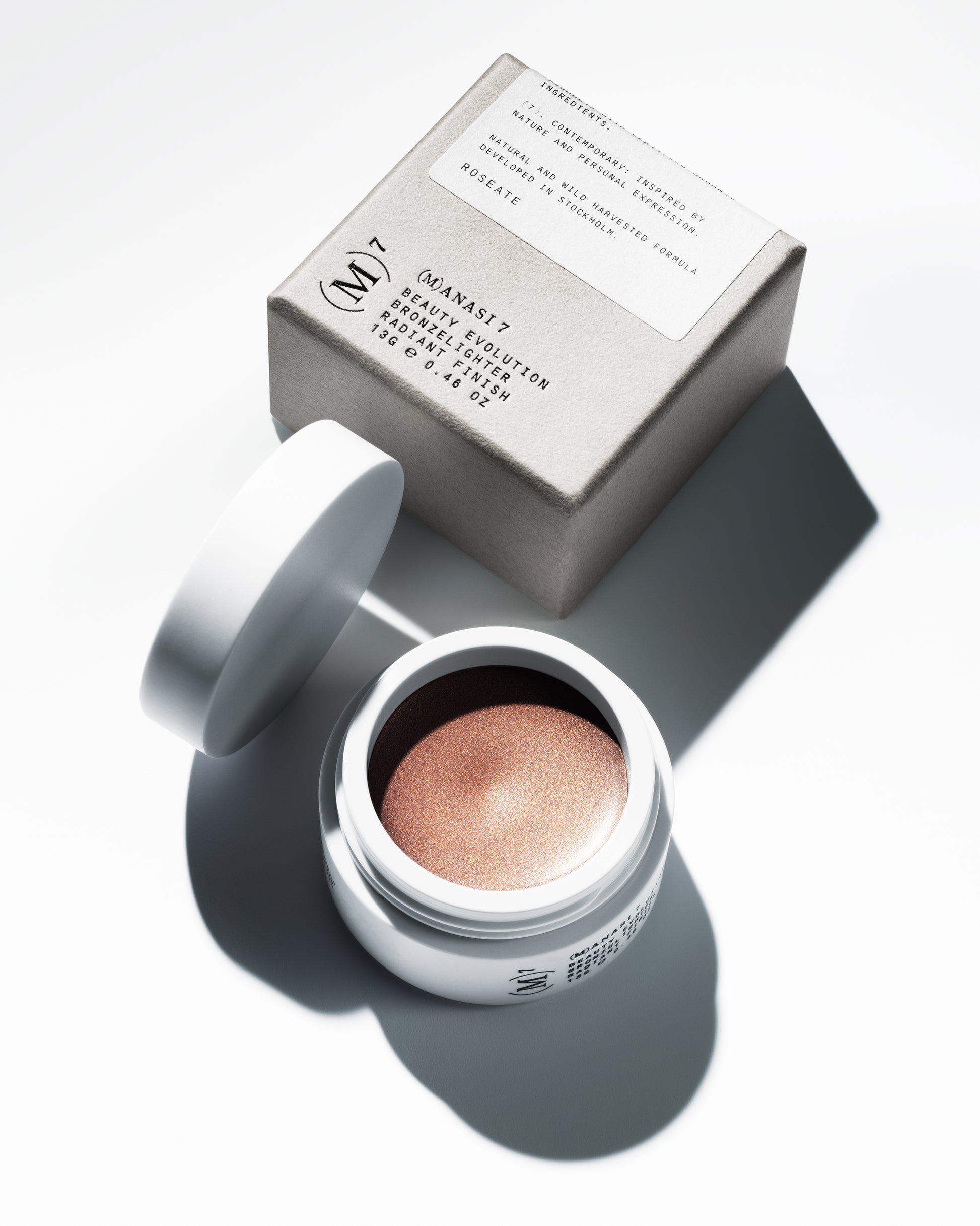 The Manasi 7 bronzelighter in the shade Roseate in a small round container and its packaging on a white background