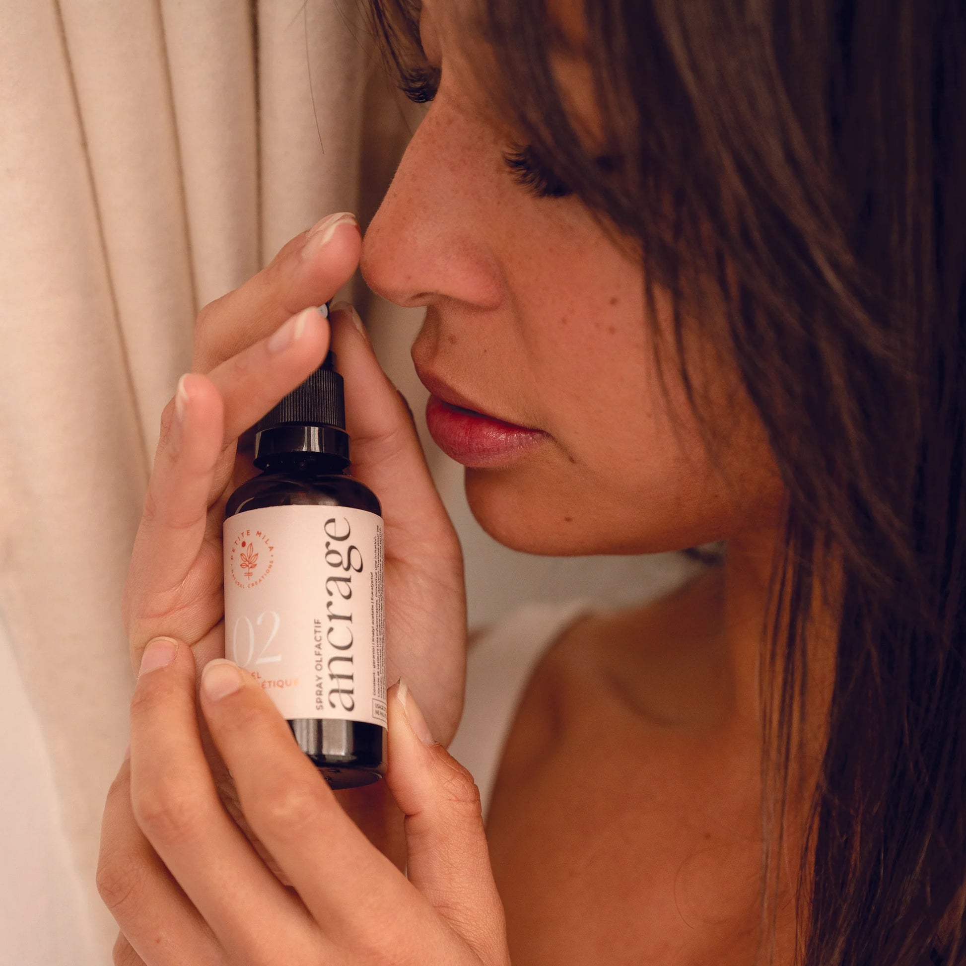 Woman holding a bottle of 'ancrage' near her nose with a neutral background