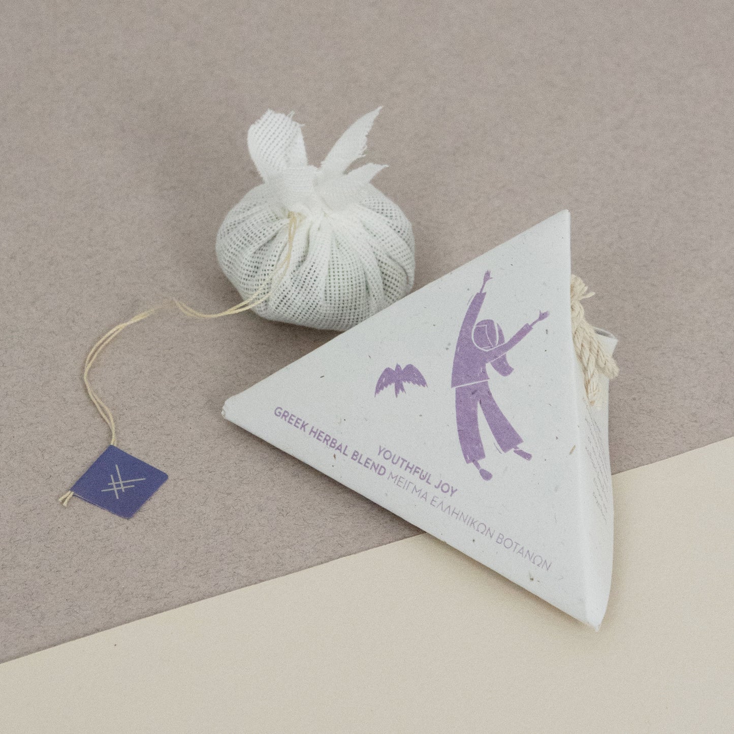 Triangular packaging and a cotton teabag of Youthful joy by Arritos with a purple design on a beige surface